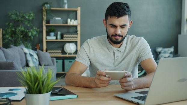 Man using smartphone at home office with laptop and plants.