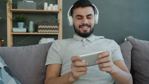Free Adult male enjoying music and gaming on smartphone wearing headphones in a cozy living room. Stock Photo