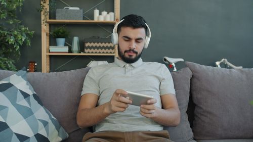 Free Young man wearing headphones and using smartphone on a couch, relaxing at home. Stock Photo