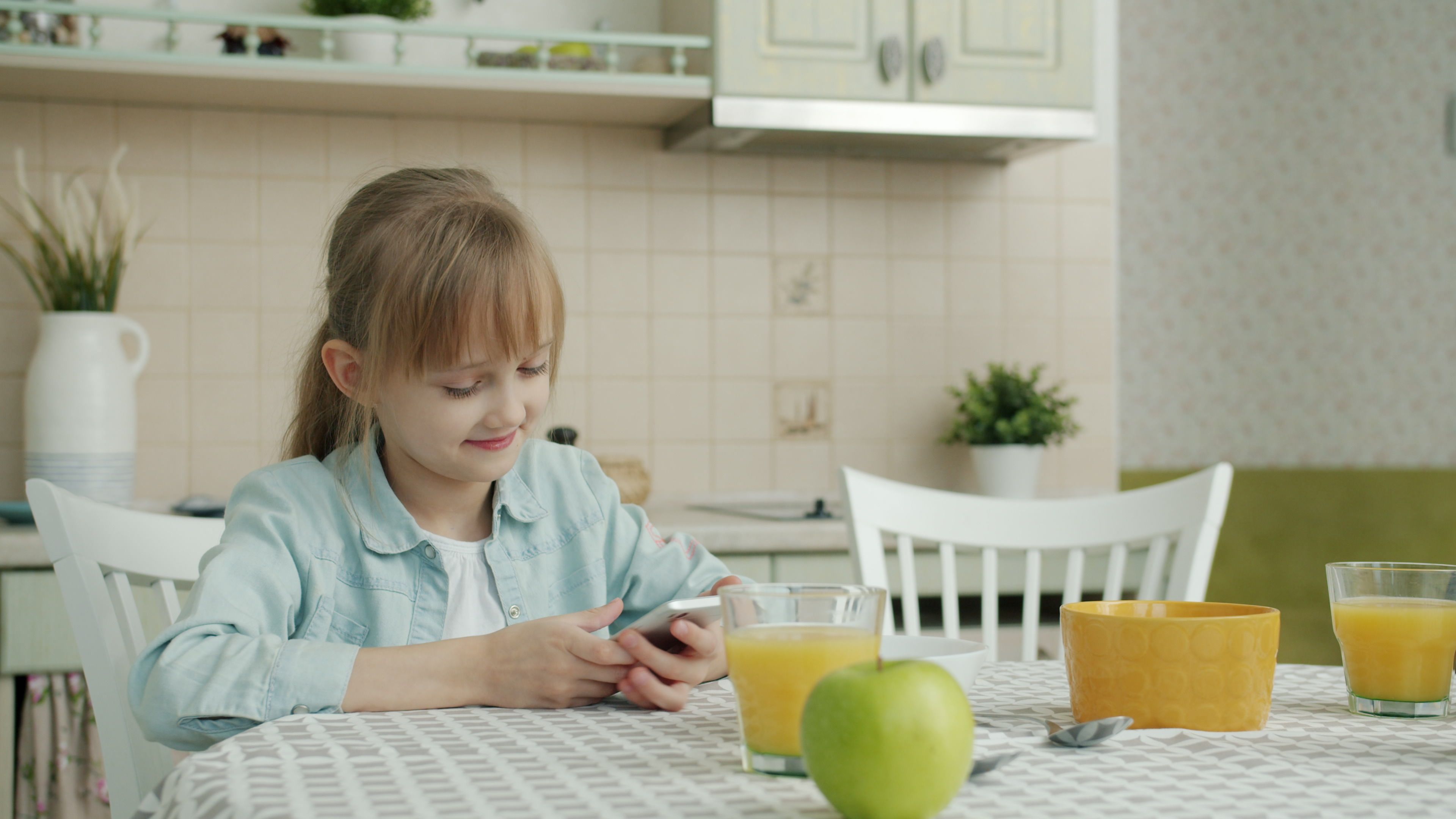 Smiling young girl uses smartphone at breakfast with juice and apple.