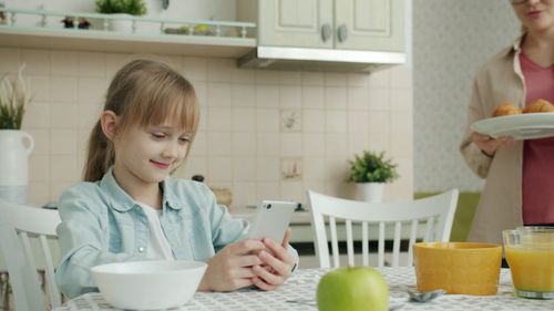 Free A young girl enjoys using a smartphone at the kitchen table. Morning routine. Stock Photo