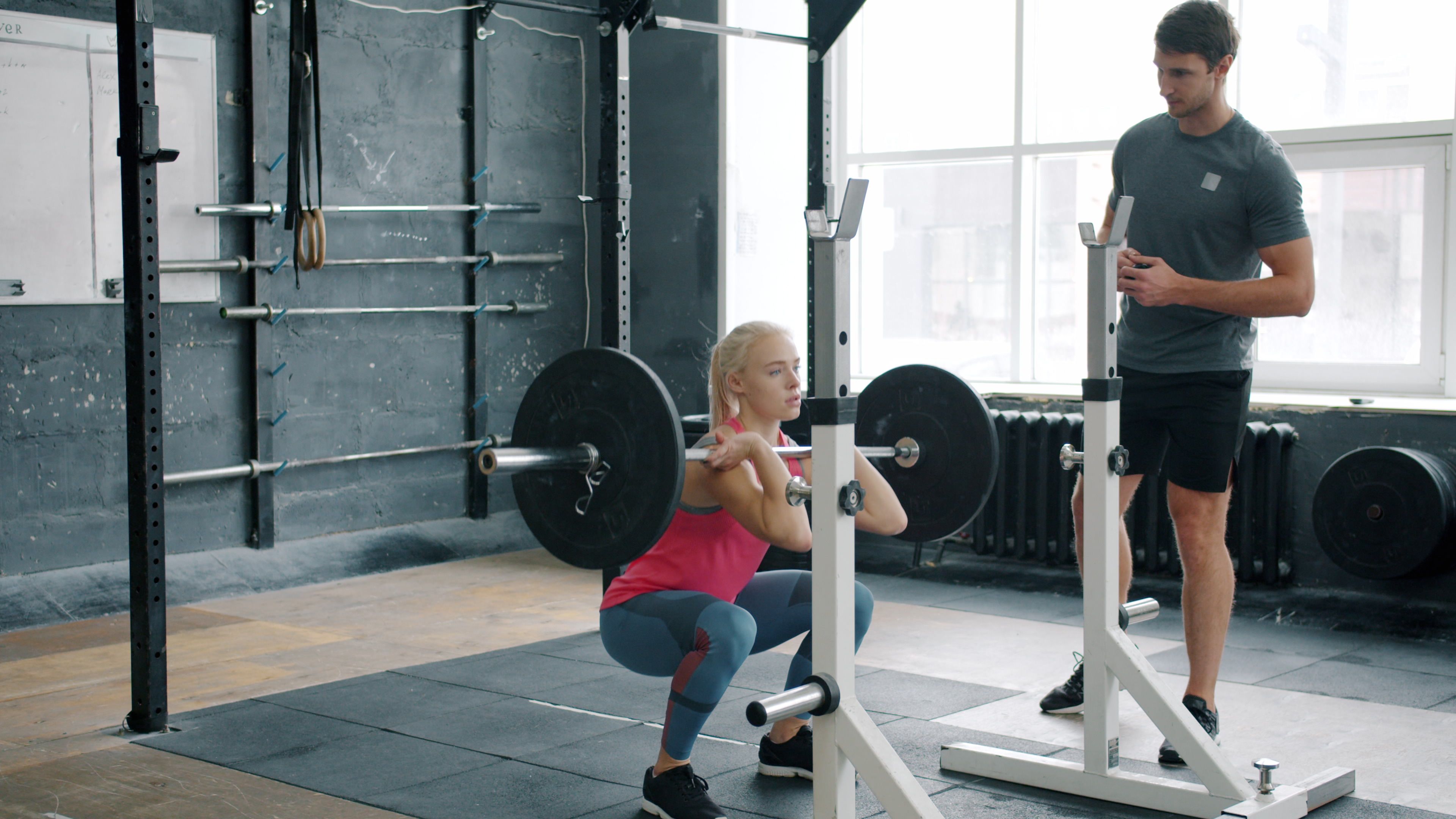 A woman performs a front squat with a barbell, guided by a trainer in a gym.