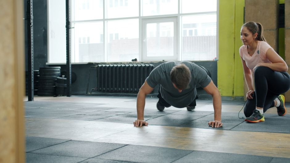 Man doing push-ups under supervision of a personal trainer in a gym setting.