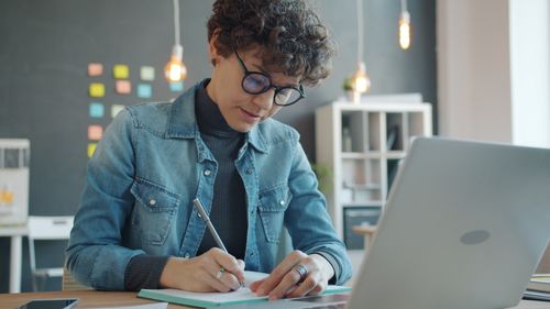 Free Woman writing notes at a desk with a laptop in a modern office setting. Stock Photo