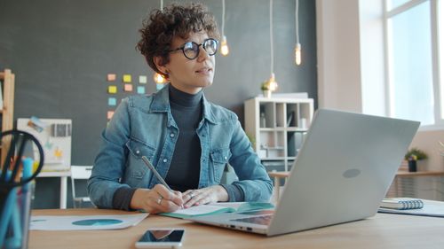 Free Young woman with glasses working at a desk with a laptop and notes. Stock Photo