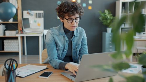 Free Focused woman in casual attire working on a laptop in a stylish office setting. Stock Photo