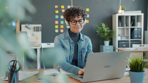 Free Young woman wearing headphones working on a laptop in a stylish modern office space. Stock Photo