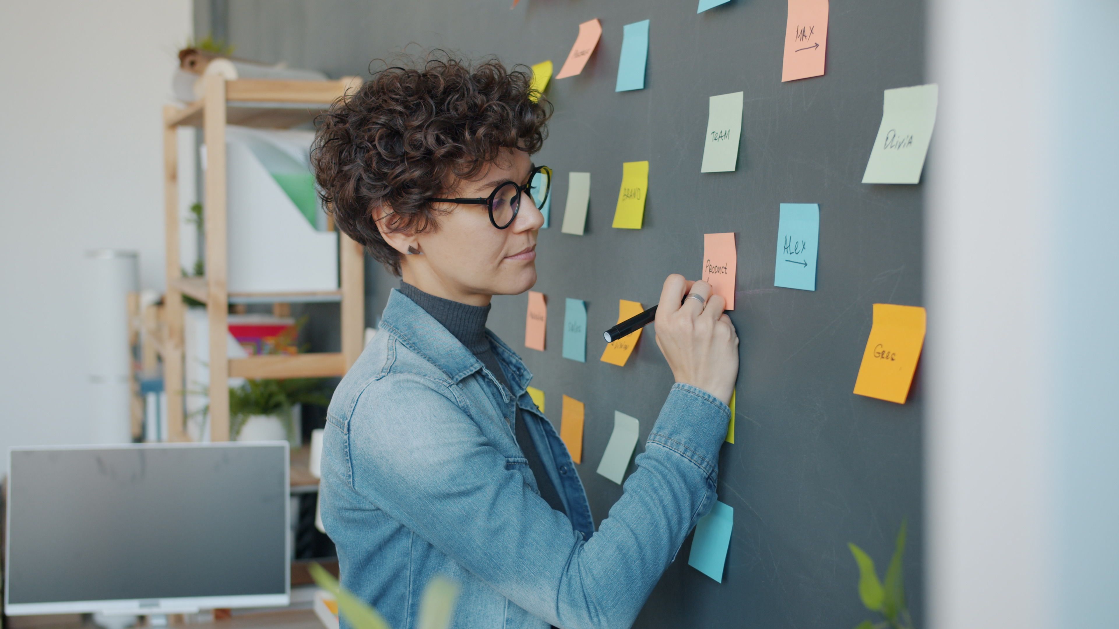 A woman in a denim jacket writes on colorful sticky notes on a blackboard in a creative office environment.