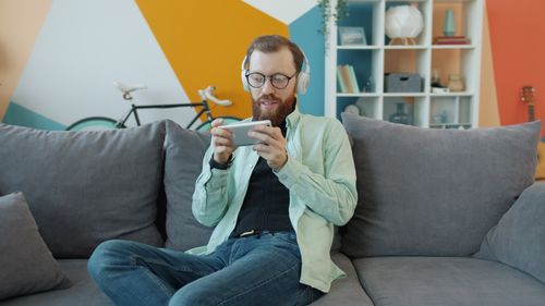 Free Man on sofa enjoying music and smartphone gaming in a modern living room setting. Stock Photo