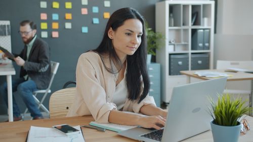 Free Woman working on laptop in a modern office with a colleague in the background. Stock Photo