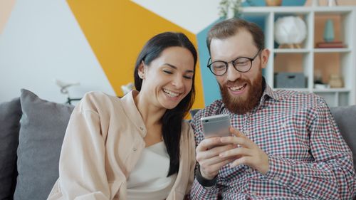 Free Smiling couple sitting on couch, sharing joyful moment while looking at smartphone screen. Stock Photo