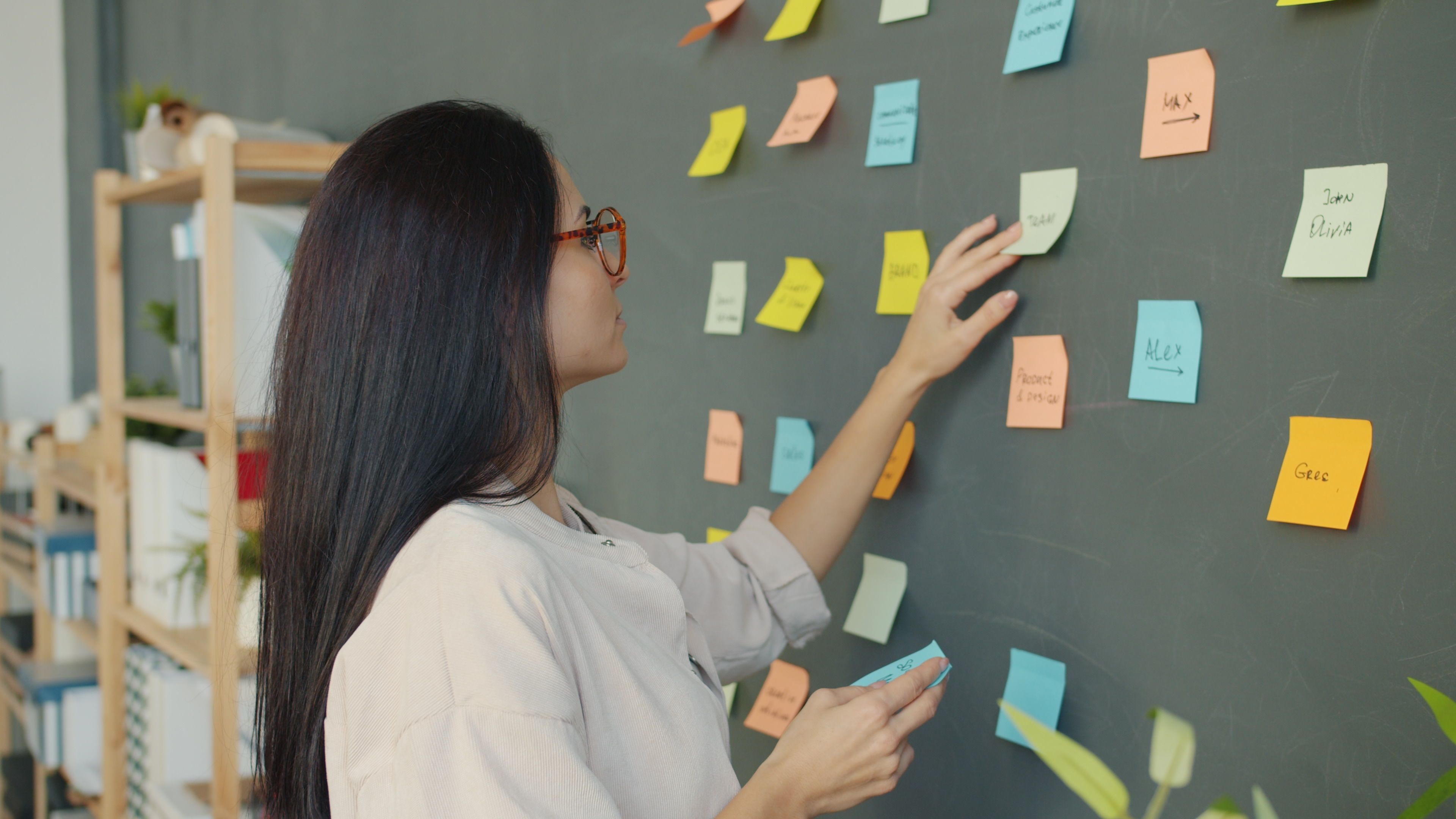 Woman placing colorful sticky notes on a blackboard in a modern office setting.