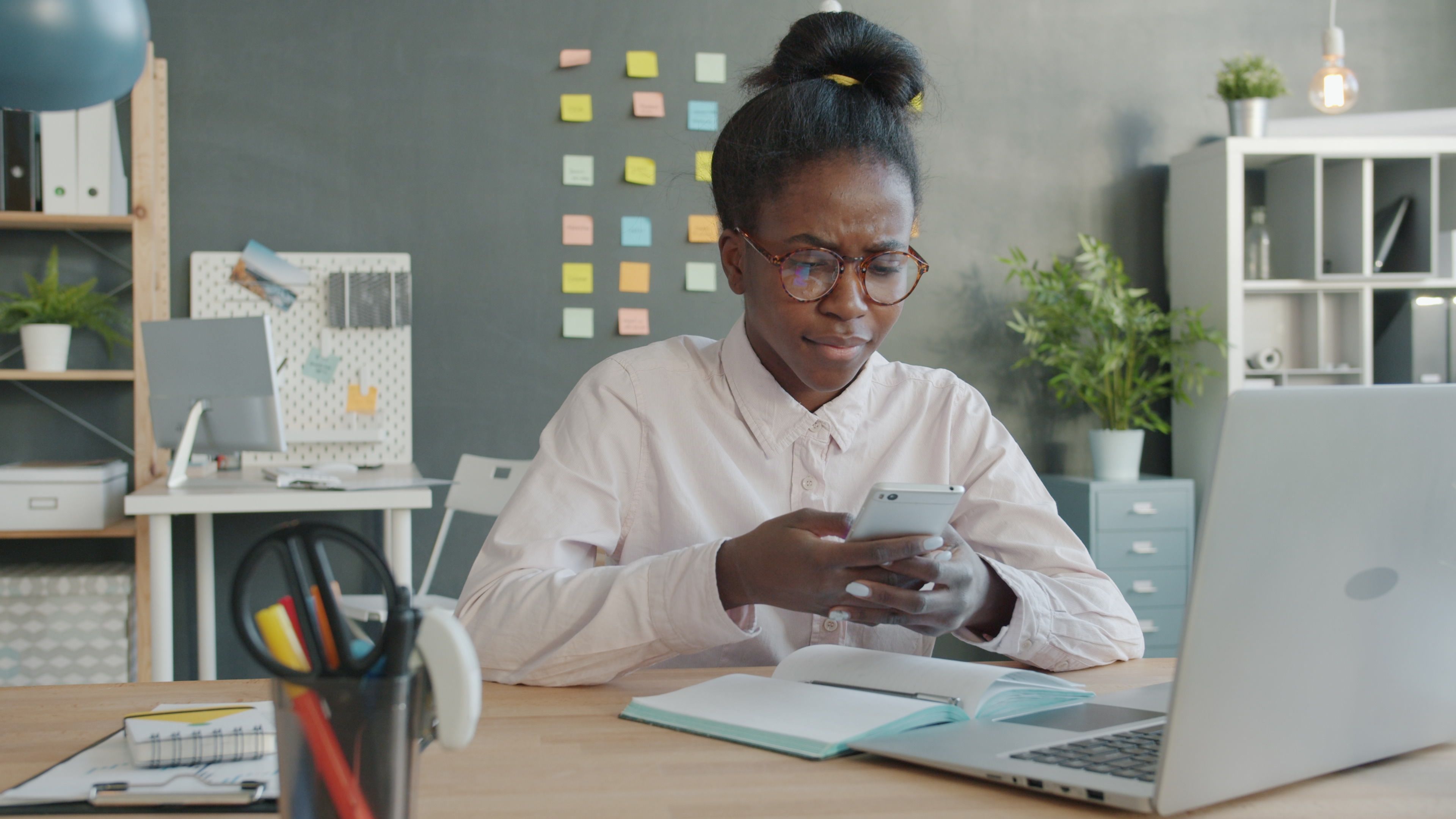 Young woman using a smartphone at her desk, surrounded by office supplies and a laptop.