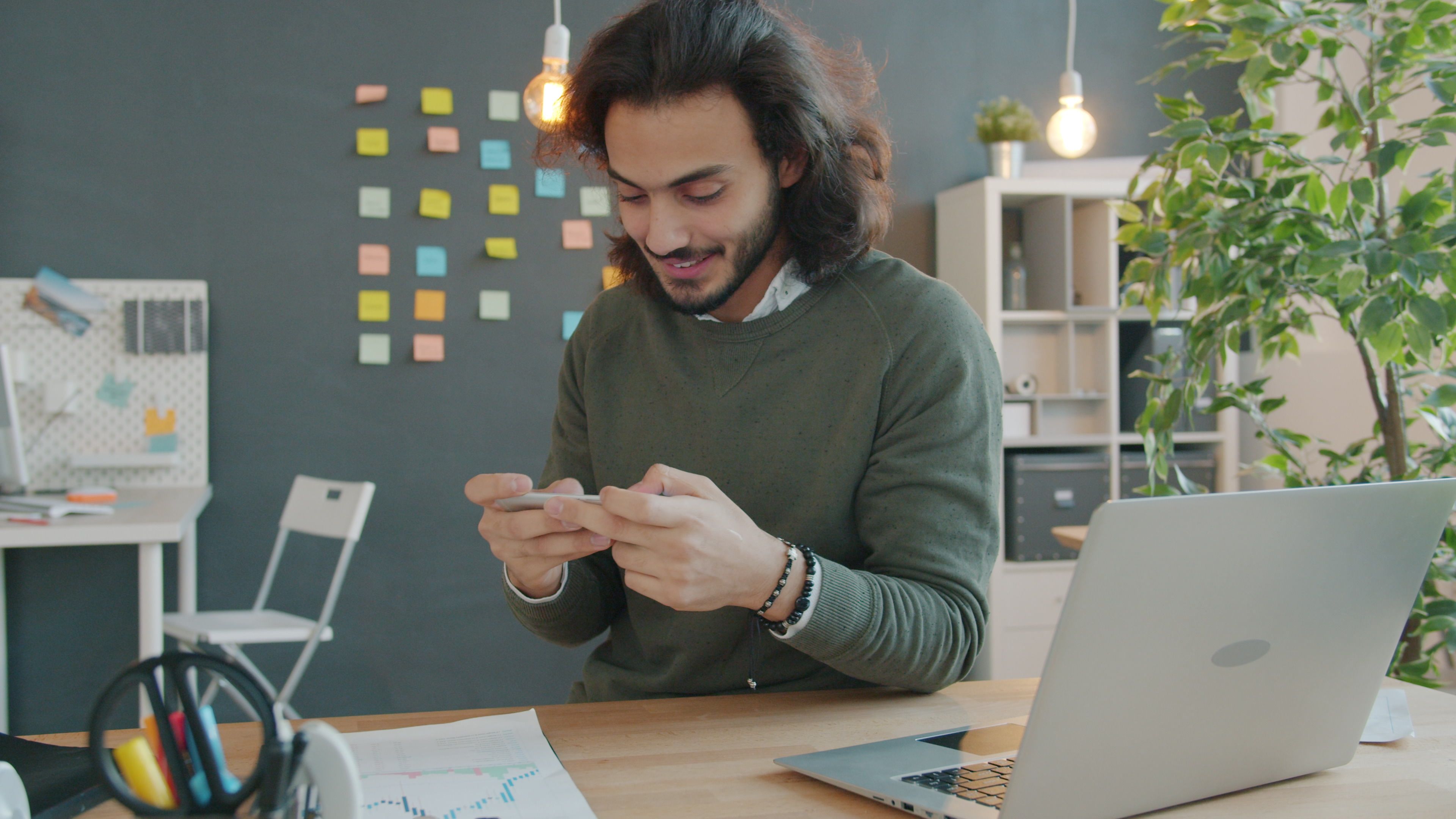 Young man using smartphone in a modern, creative office with sticky notes and a laptop.
