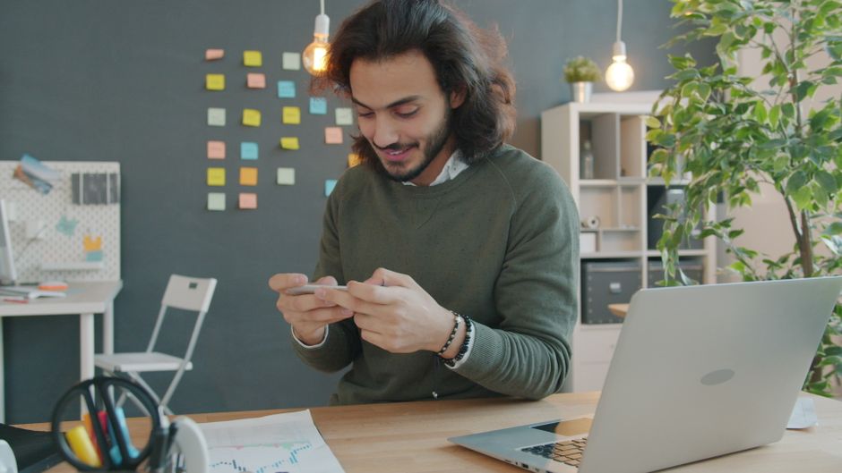 Young man using smartphone in a modern, creative office with sticky notes and a laptop. Young man using smartphone in a modern, creative office with sticky notes and a laptop.