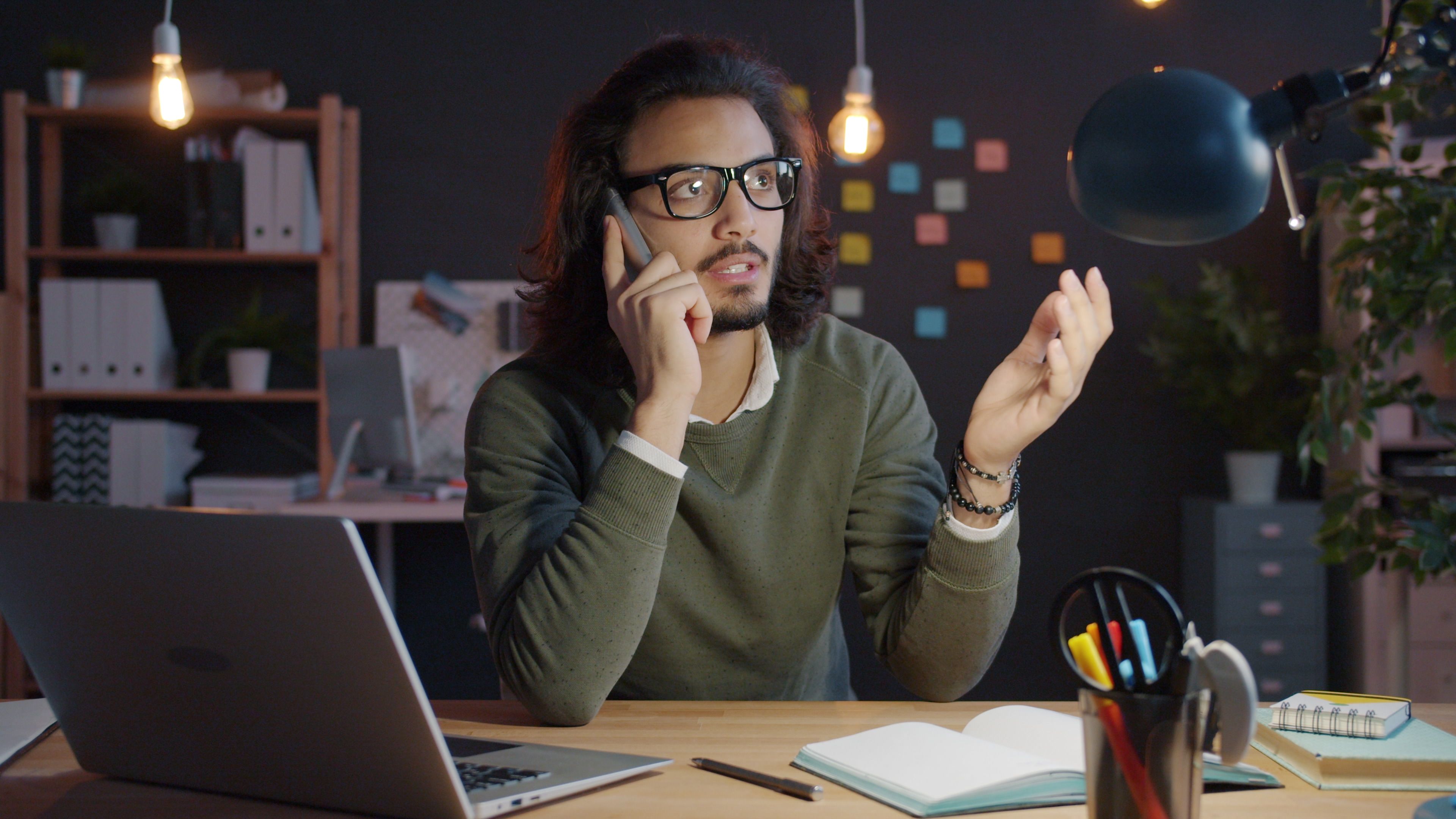Man talking on phone at home office desk with laptop and notes, looking focused.