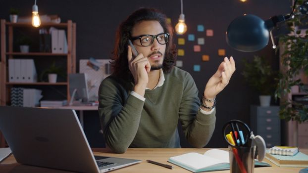 Man talking on phone at home office desk with laptop and notes, looking focused.