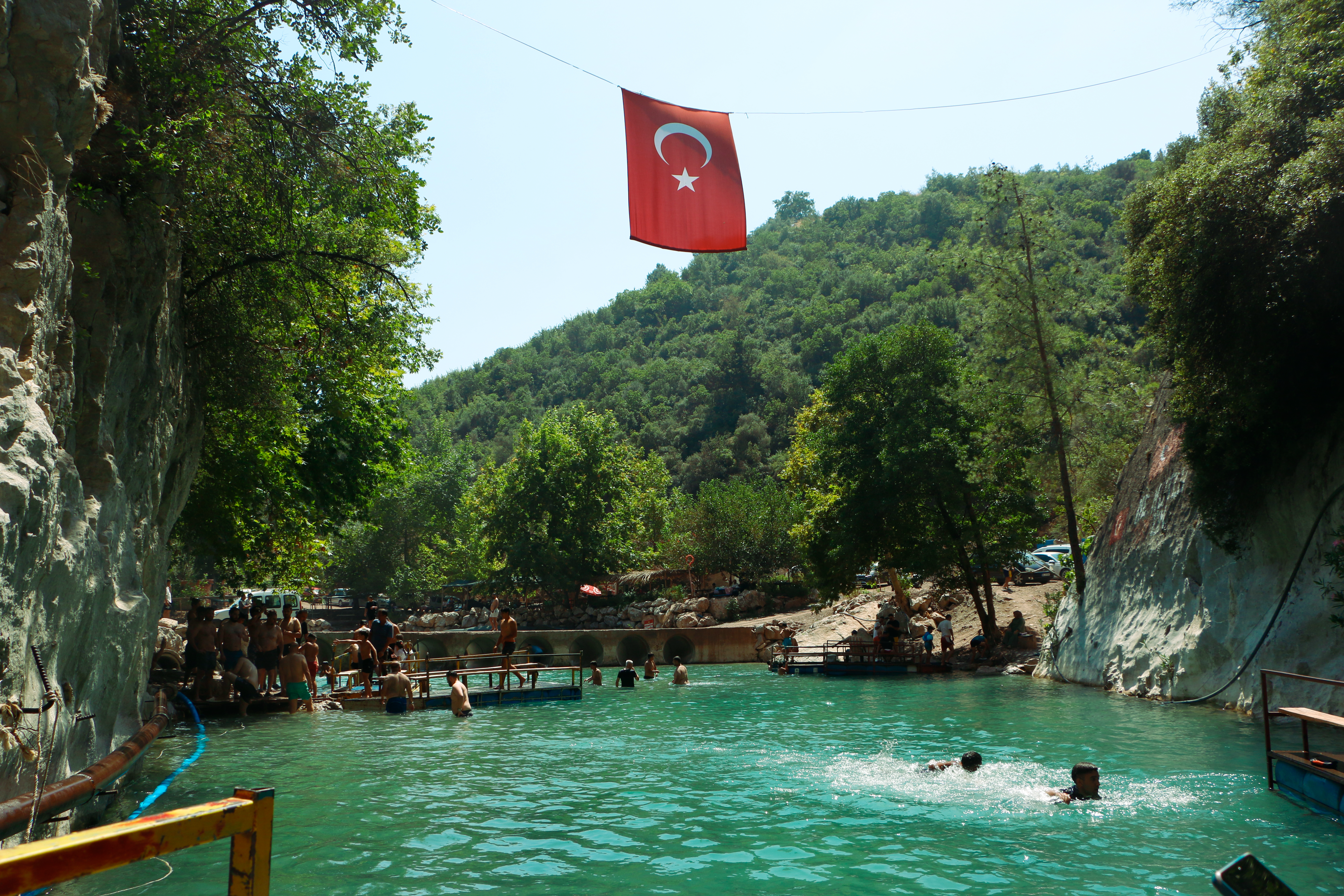 People enjoying a scenic natural swimming area in Mersin, Türkiye under a Turkish flag.