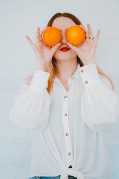 一位穿着白色衬衫的女人，在极简主义的背景下，俏皮地把橘子举到眼前