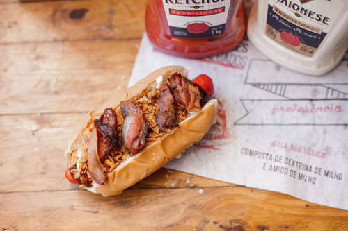 Free Appetizing hot dog topped with bacon, crispy onions, and condiments on a wooden table. Stock Photo