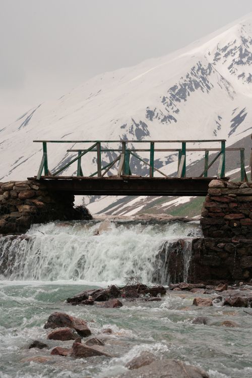 Free A rustic wooden bridge spans a turbulent stream against a backdrop of snow-covered mountains. Stock Photo