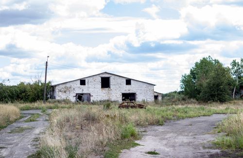 Free Old abandoned warehouse in a rural area with overgrown grass and cloudy skies. Stock Photo