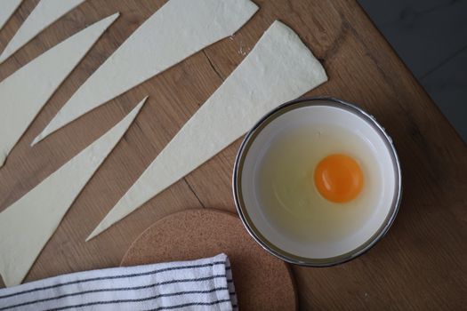 Top view of pastry dough slices and a raw egg in a bowl on a wooden table.