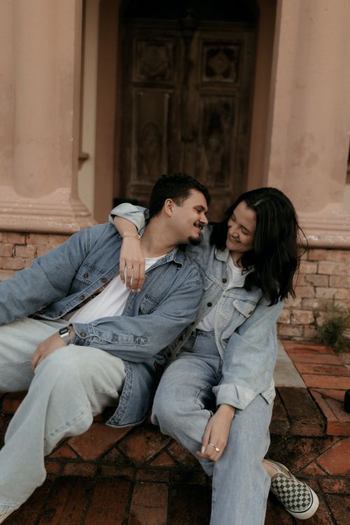 Free A couple in denim sitting close on brick steps outside an old building, smiling at each other. Stock Photo