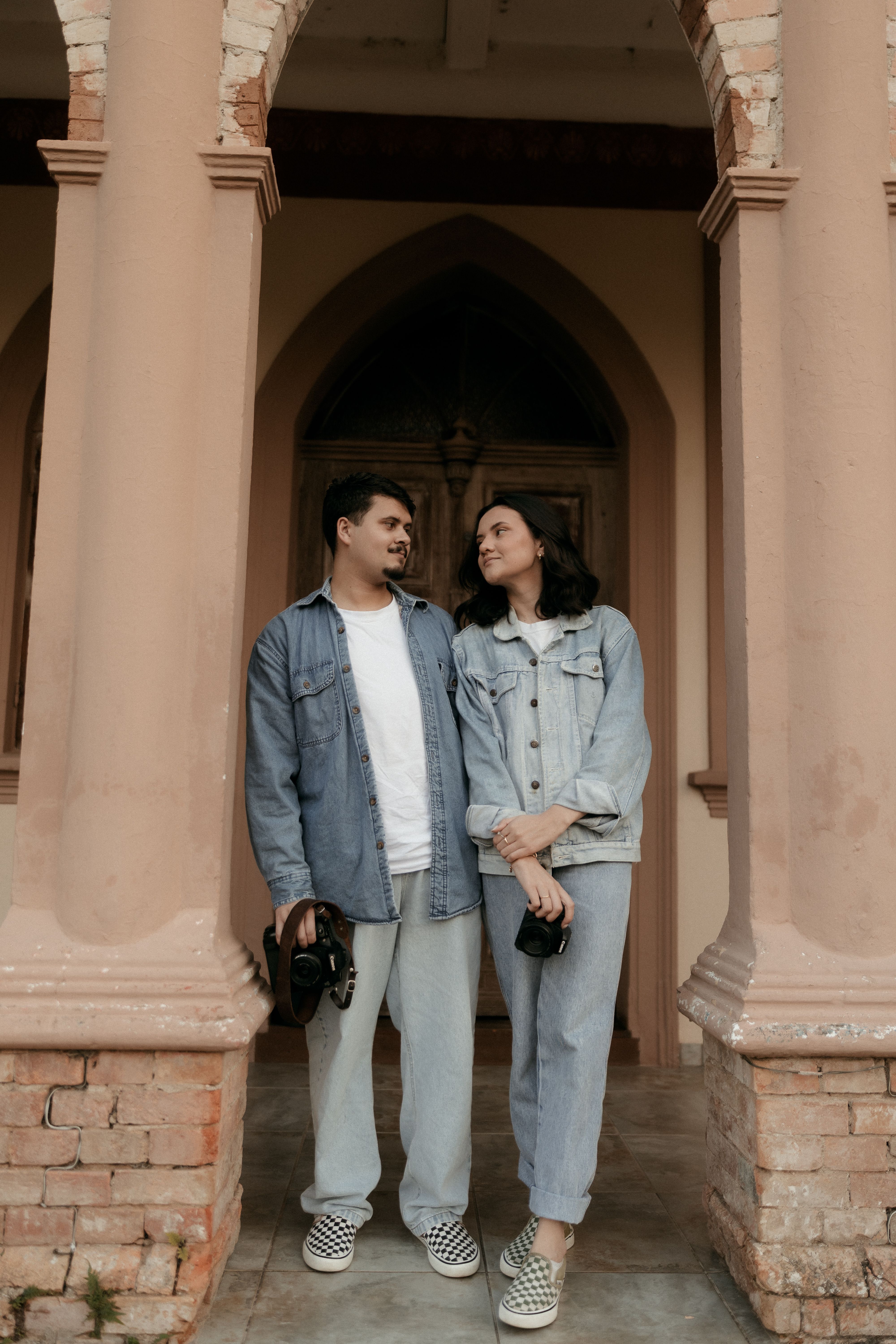 A couple holding cameras, dressed in denim, stand romantically in front of an arched vintage building.