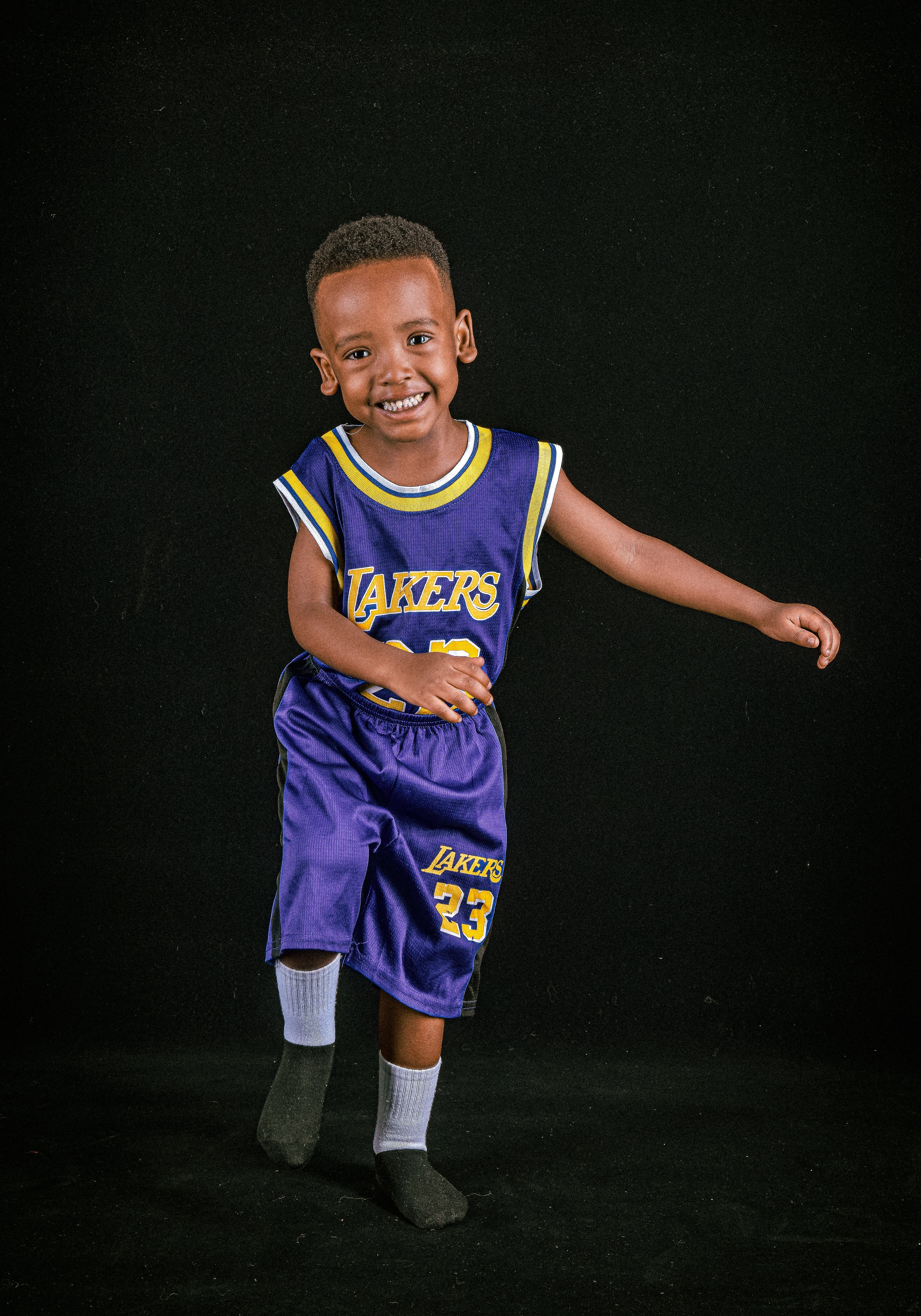 A joyful child wearing a basketball jersey smiles for the camera on a dark backdrop.