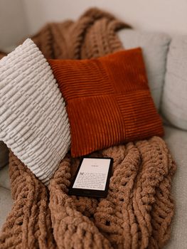 A warm and inviting setup featuring pillows, a chunky knit blanket, and an e-reader, perfect for relaxation.