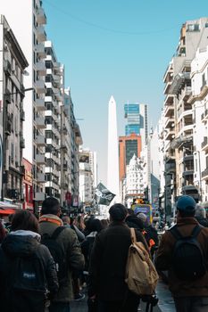 Busy urban street scene with people facing an iconic obelisk under clear skies.