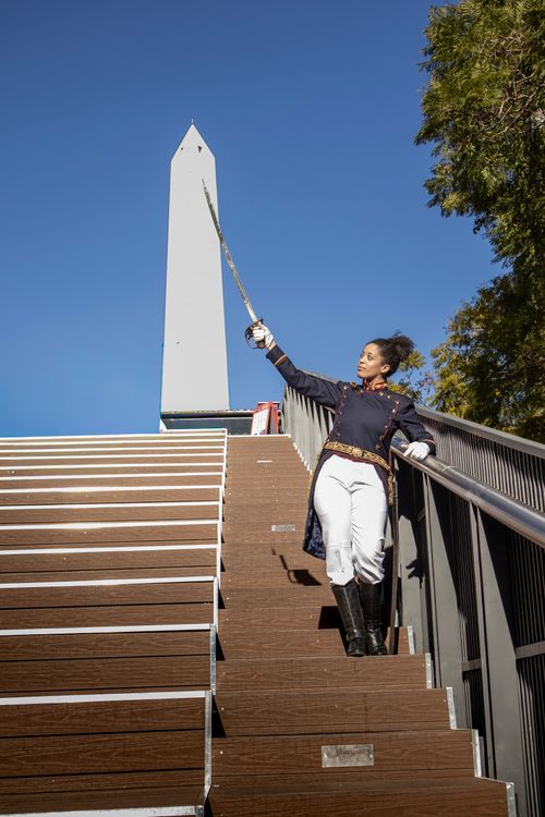 Free A confident performer in uniform holding a sword on steps near an obelisk under a clear blue sky. Stock Photo