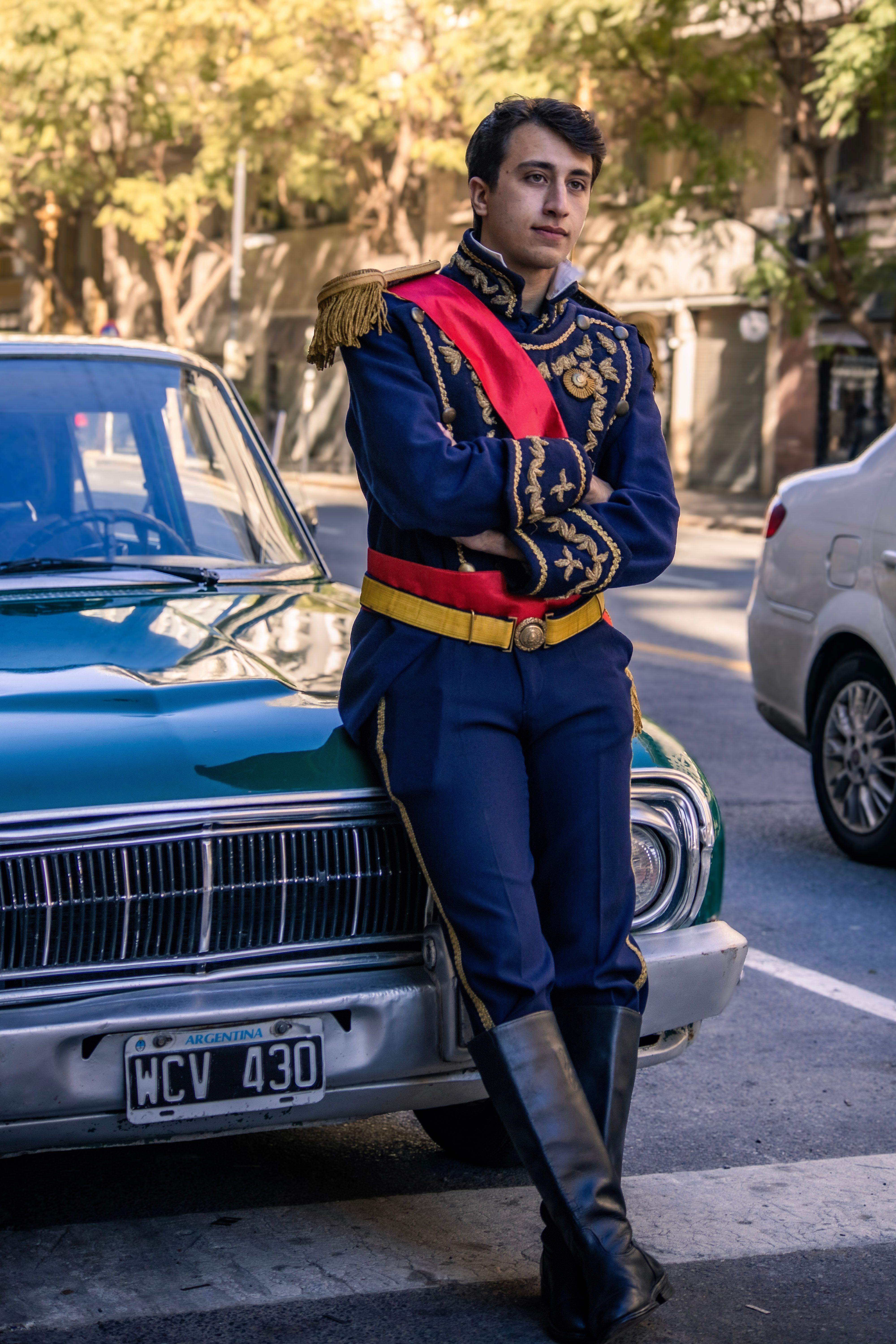 A man in historical military attire leans on a vintage car in a city setting.