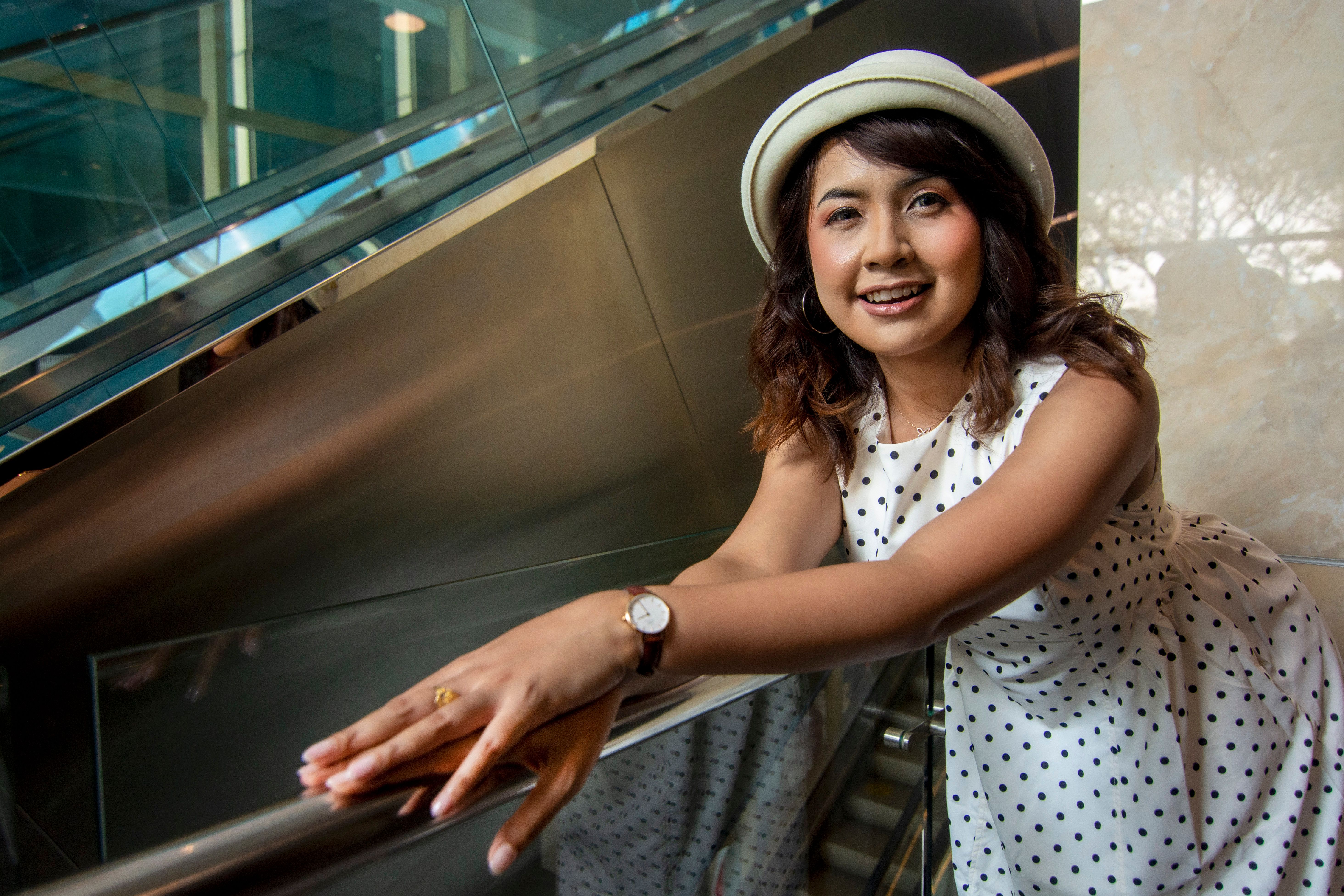 Smiling woman in polka dot dress and hat on an escalator indoors in Jakarta.