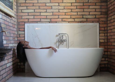 A woman relaxes in a modern bathtub in a brick-walled bathroom.