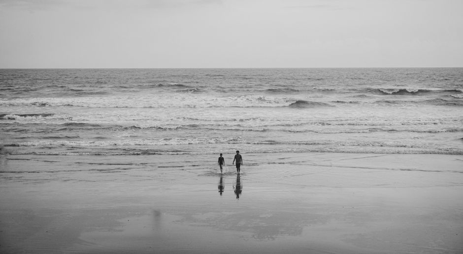 Black and white photo of two people walking on Morjim beach, India, with ocean waves.