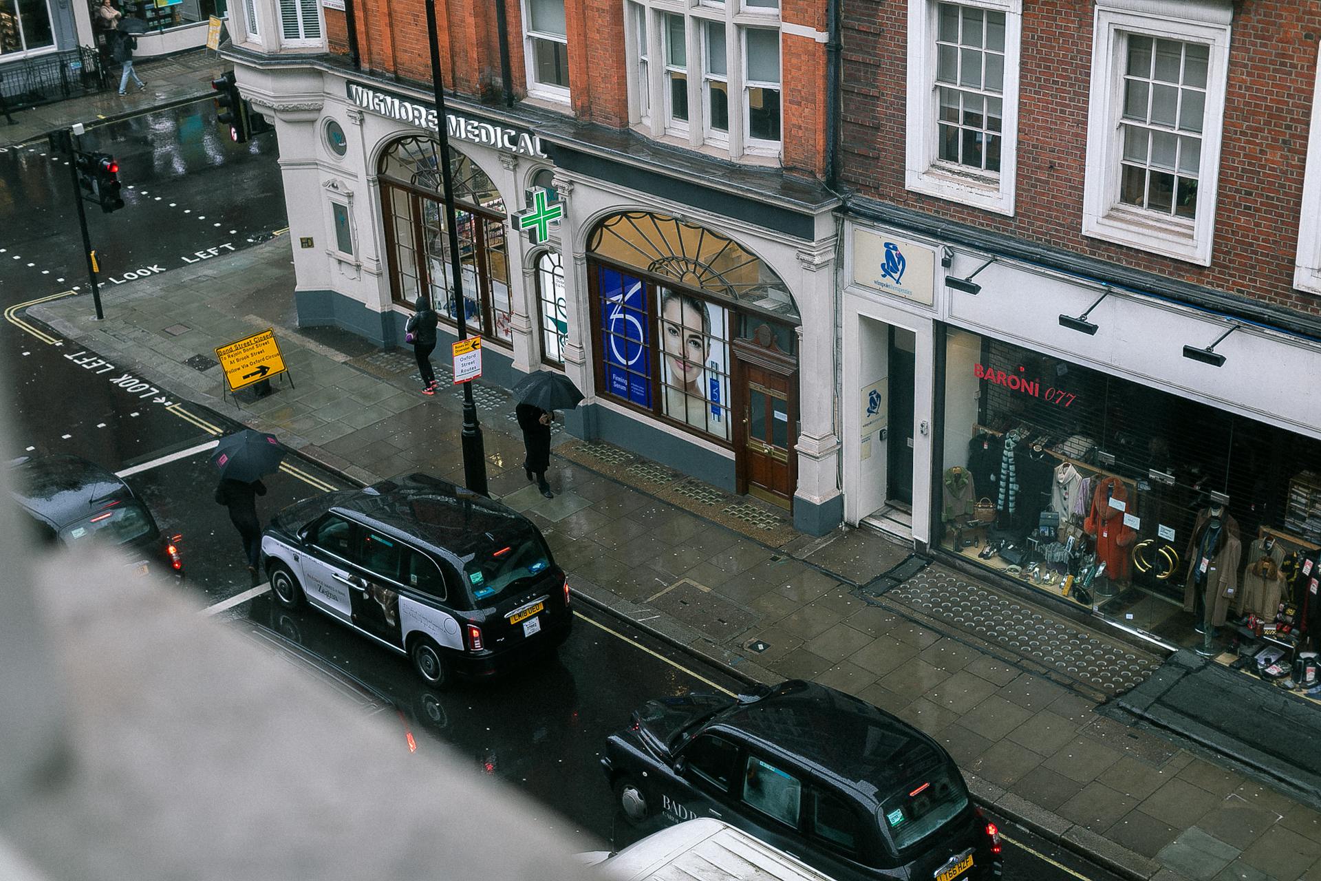 Overhead view of a rainy street in London with black cabs and pedestrians under umbrellas.