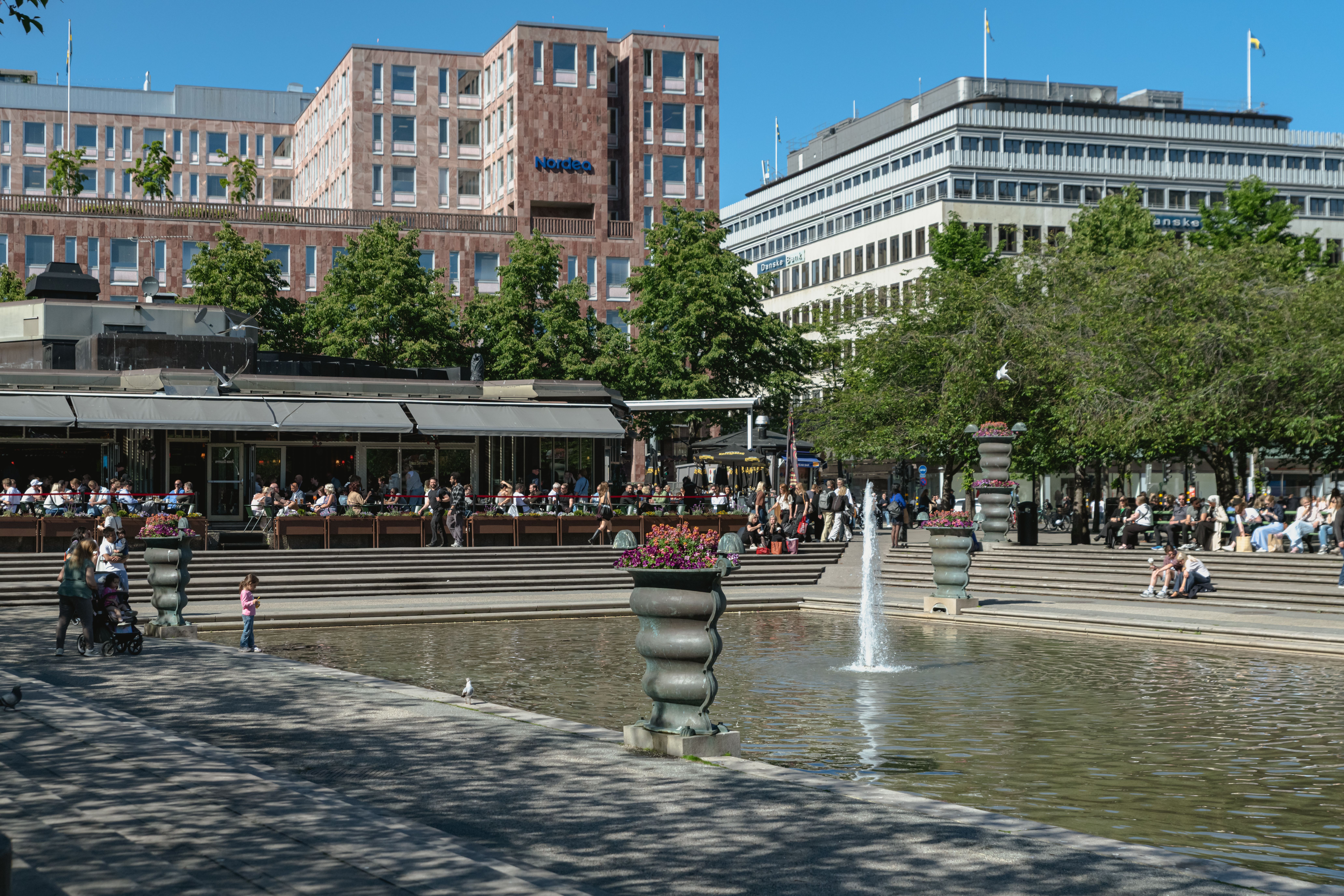People enjoying a sunny day at an urban park with fountain and seating in Stockholm.