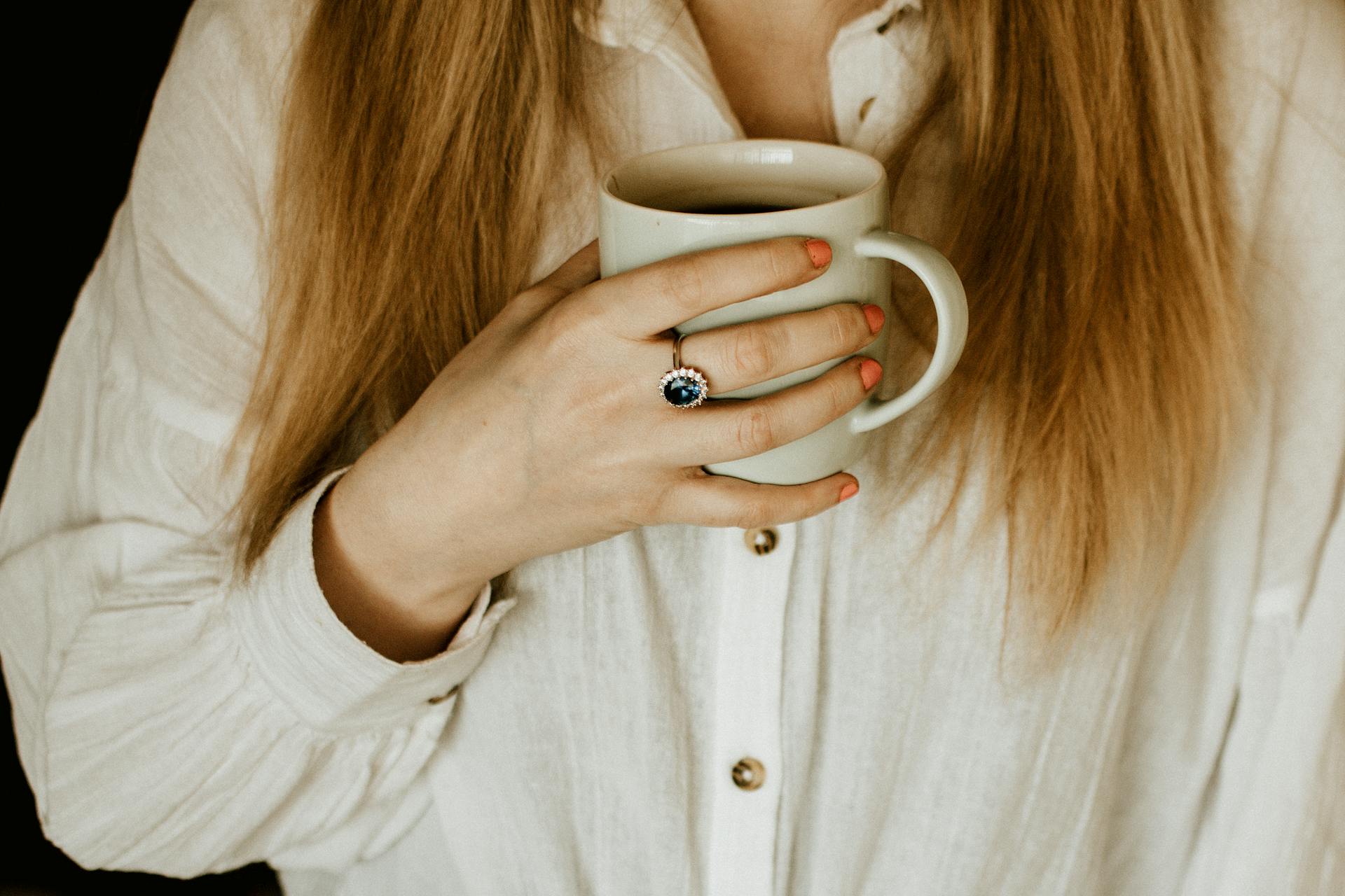 A close-up shot of a woman holding a coffee mug, wearing a ring with a blue gemstone.