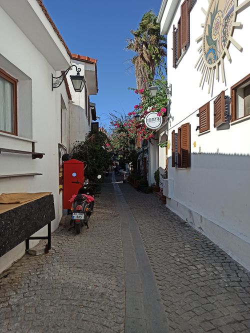 Free A scenic narrow alley with sunlight and charming European architecture, featuring lush plants and a scooter. Stock Photo