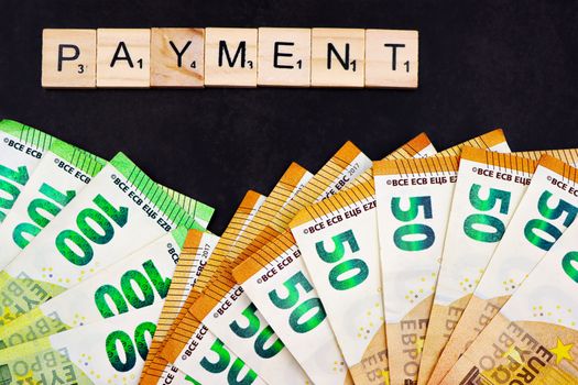 Euro banknotes fanned out with 'PAYMENT' spelled in Scrabble tiles, representing finance and commerce.