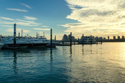 Sunset view of yachts moored at a marina with a city skyline backdrop.