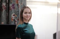 一位身穿绿色连衣裙的美丽女子在室内微笑，四周环绕着花卉窗帘