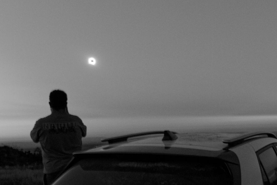 Silhouette en noir et blanc d'une personne observant une éclipse solaire, près d'une voiture
