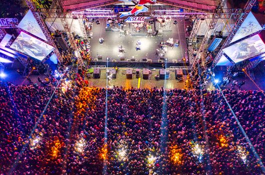A drone-captured aerial view of a colorful crowd enjoying a live concert at night with bright stage lights.