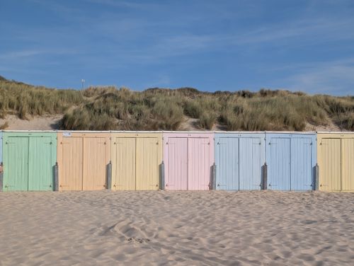 Free A row of pastel-colored beach huts along a sandy beach with grassy dunes under a clear blue sky. Stock Photo