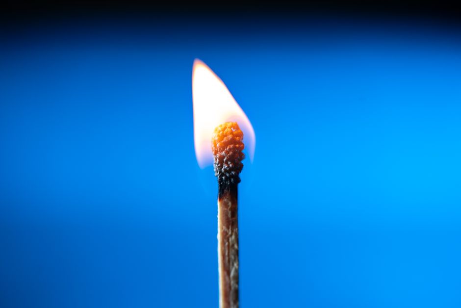 A close-up image of a single matchstick burning, casting a warm glow against a blue background.