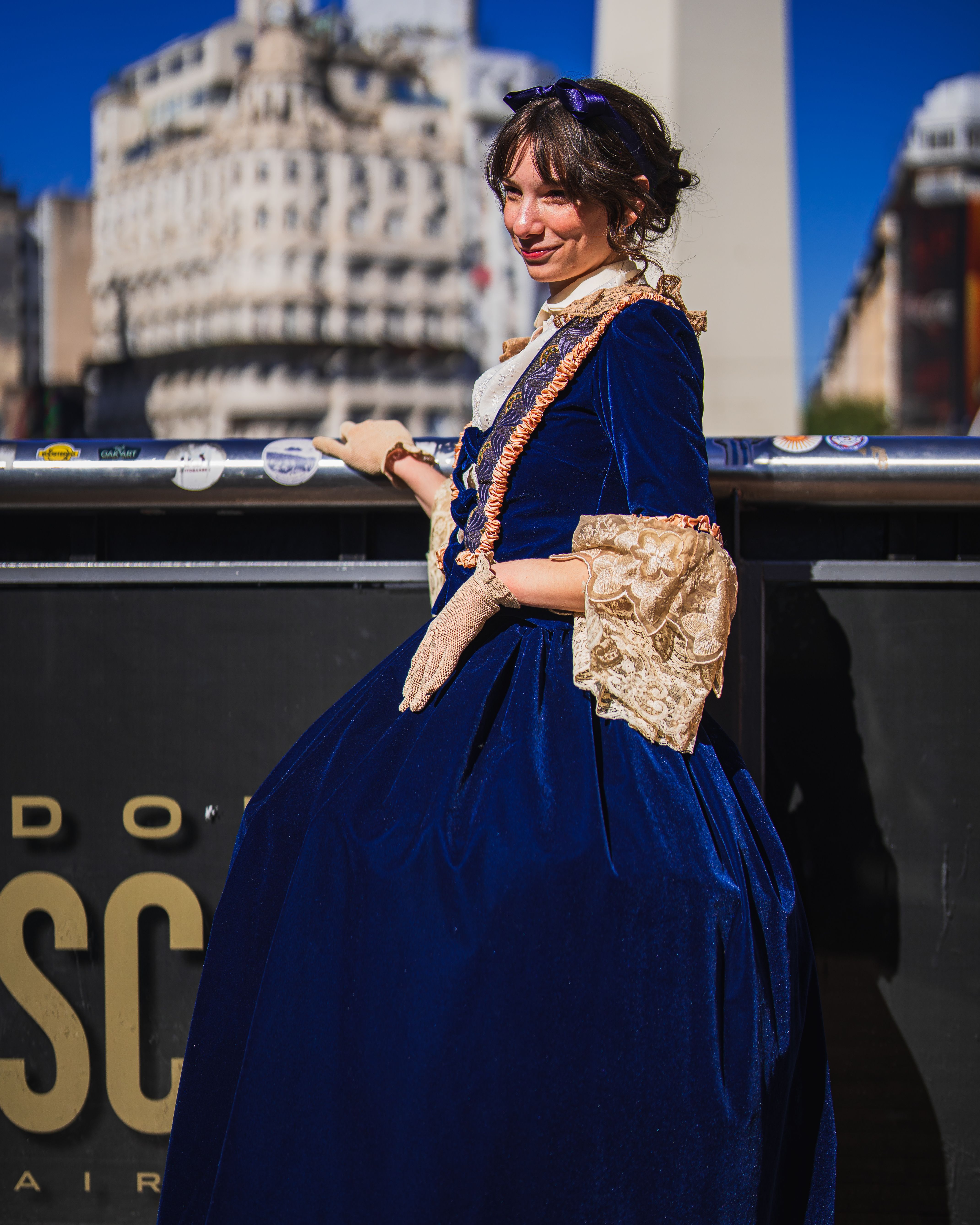 Woman in vintage blue gown poses outdoors with city skyline backdrop.