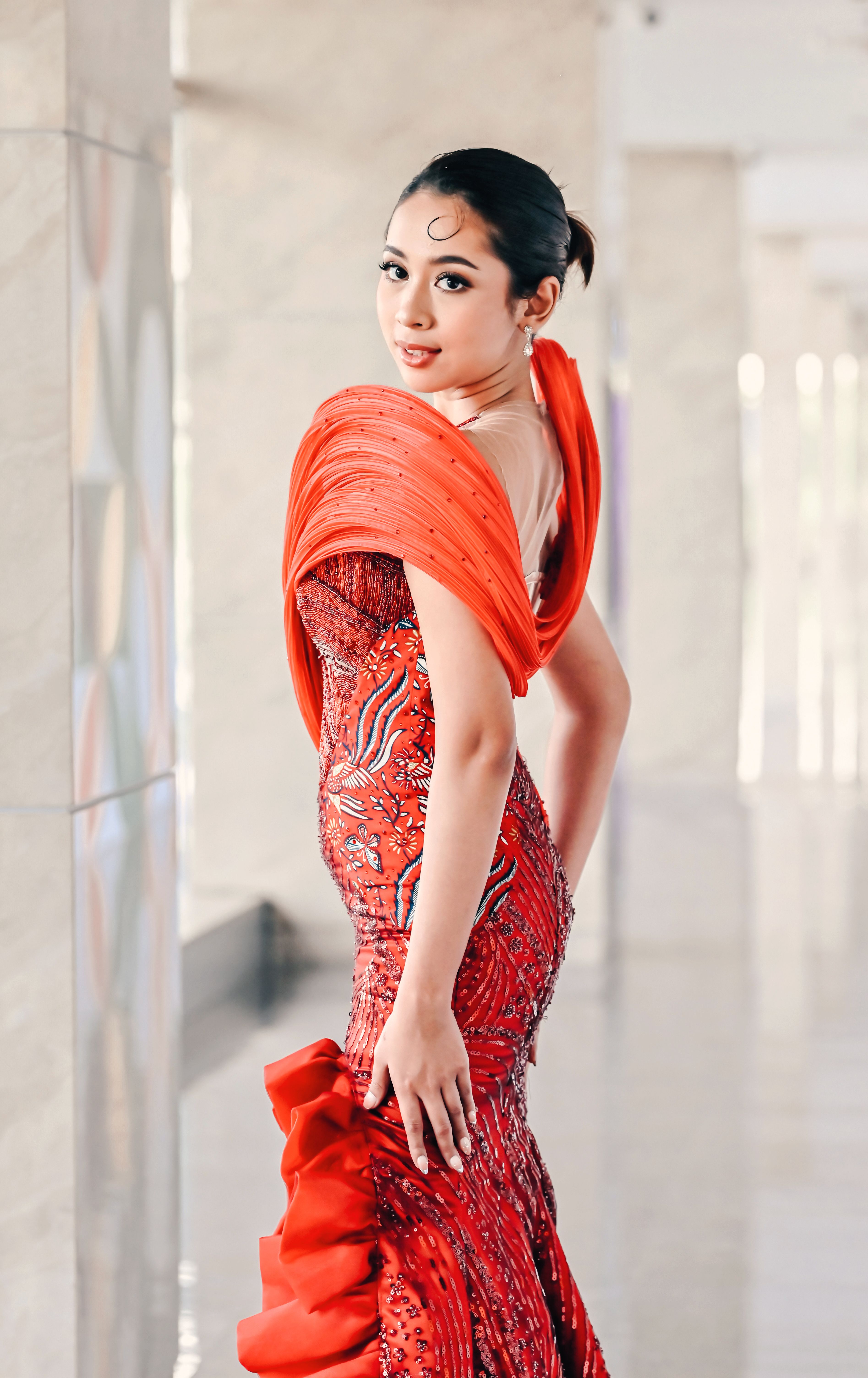 Stylish woman in an intricately designed red dress poses indoors, exuding elegance and confidence.
